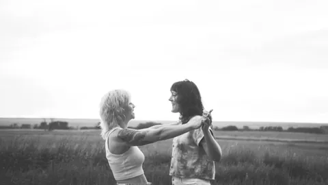 Black and white photo of two women smiling and holding hands in a grassy field under an overcast sky.