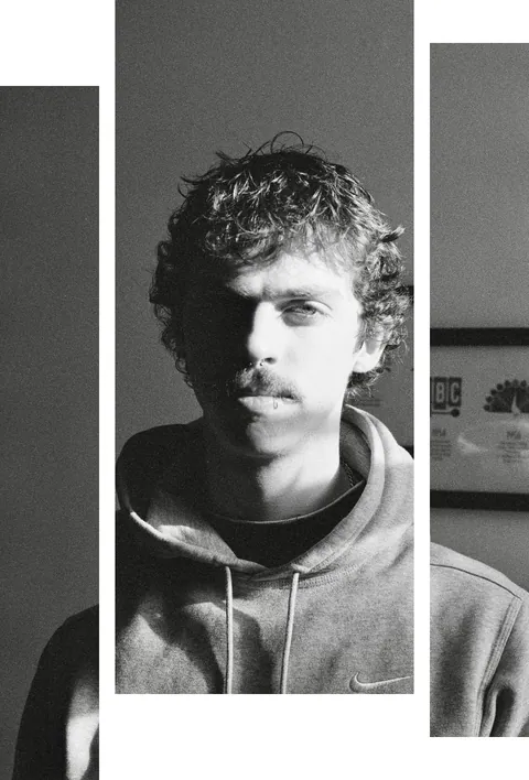 Black and white portrait of a curly-haired young man with a mustache in a hoodie, with a framed plaque behind him.