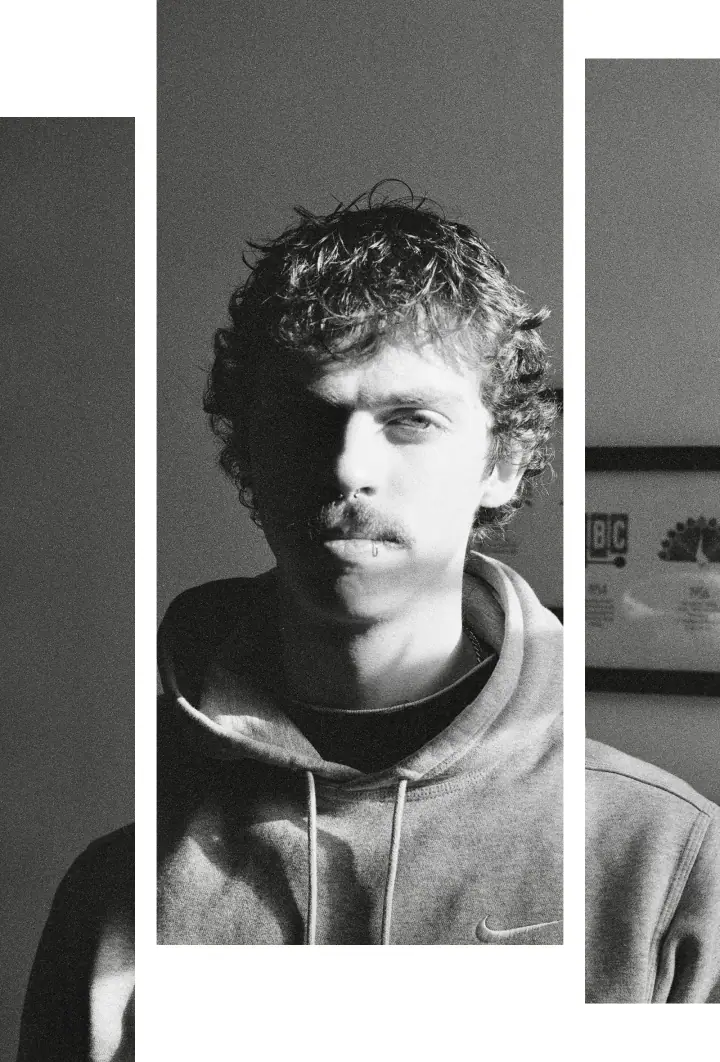 Black and white portrait of a curly-haired young man with a mustache in a hoodie, with a framed plaque behind him.