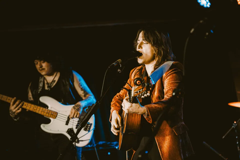 Singer in a rust velvet jacket plays acoustic guitar at a mic as a bassist with a white bass performs behind.
