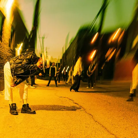 Person in a black leather jacket and white pants bends over on a glowing orange night alley, others blurred behind.